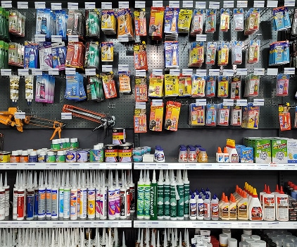 Rayons d'un magasin avec une vaste sélection de colles, mastics et pistolets à calfeutrer. Nombreux produits UHU, SIKA, BONY visibles.