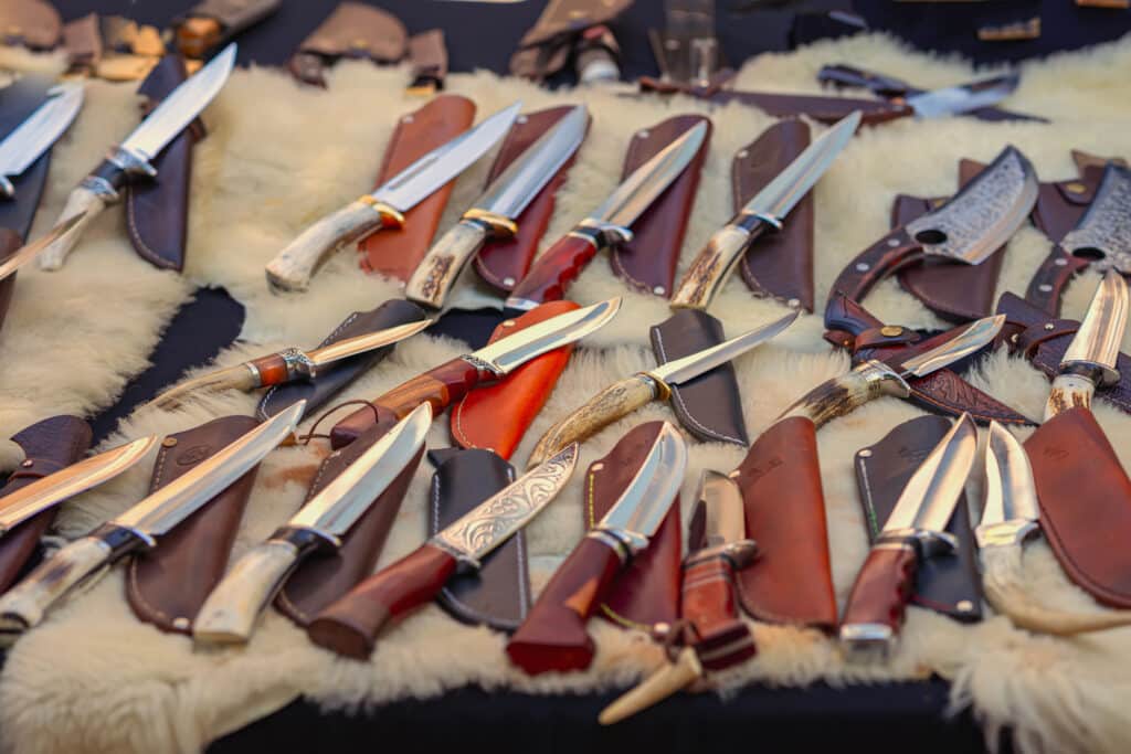 Gros plan sur une collection de couteaux de chasse artisanaux avec manches en bois et corne, dans des étuis en cuir, exposés sur une fourrure claire.