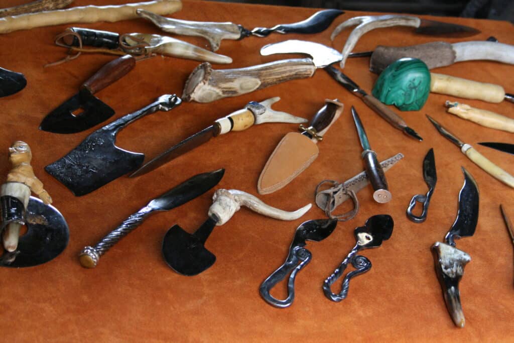Divers couteaux et outils artisanaux forgés main avec manches en bois, corne et os, disposés sur un fond marron. Inclut une pierre de malachite verte.
