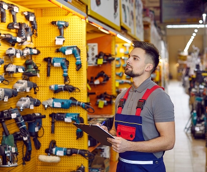 Homme en salopette vérifiant des perceuses et meuleuses sur un mur d'outils jaune vif en magasin. Il note sur un presse-papiers.