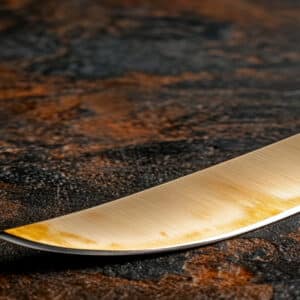 Lame de couteau dorée avec reflets, légèrement usée, posée sur un fond abstrait texturé sombre aux teintes rouille.