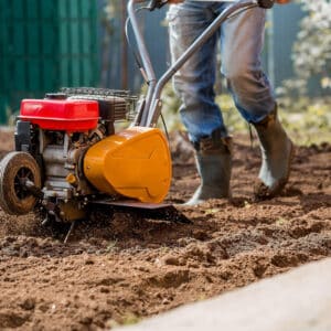 Un jardinier en jeans et bottes utilise un motoculteur rouge et jaune, mélangeant la terre pour la préparation du sol.