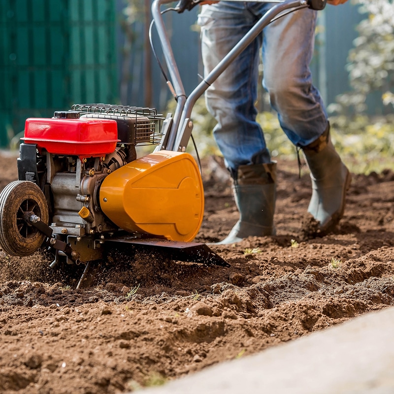 Un jardinier en jeans et bottes utilise un motoculteur rouge et jaune, mélangeant la terre pour la préparation du sol.