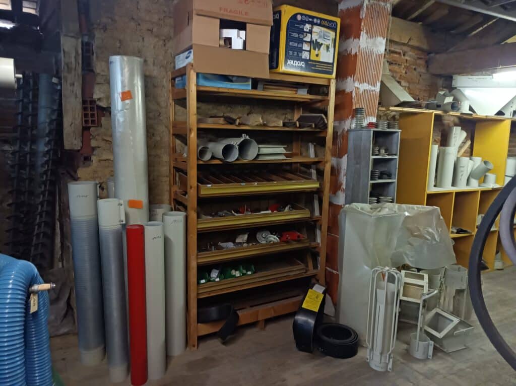 Entrepôt de stockage avec étagères en bois, tuyaux, rouleaux, et matériel industriel. Boîtes "FRAGILE" et "Brother DCP-7010" visibles.