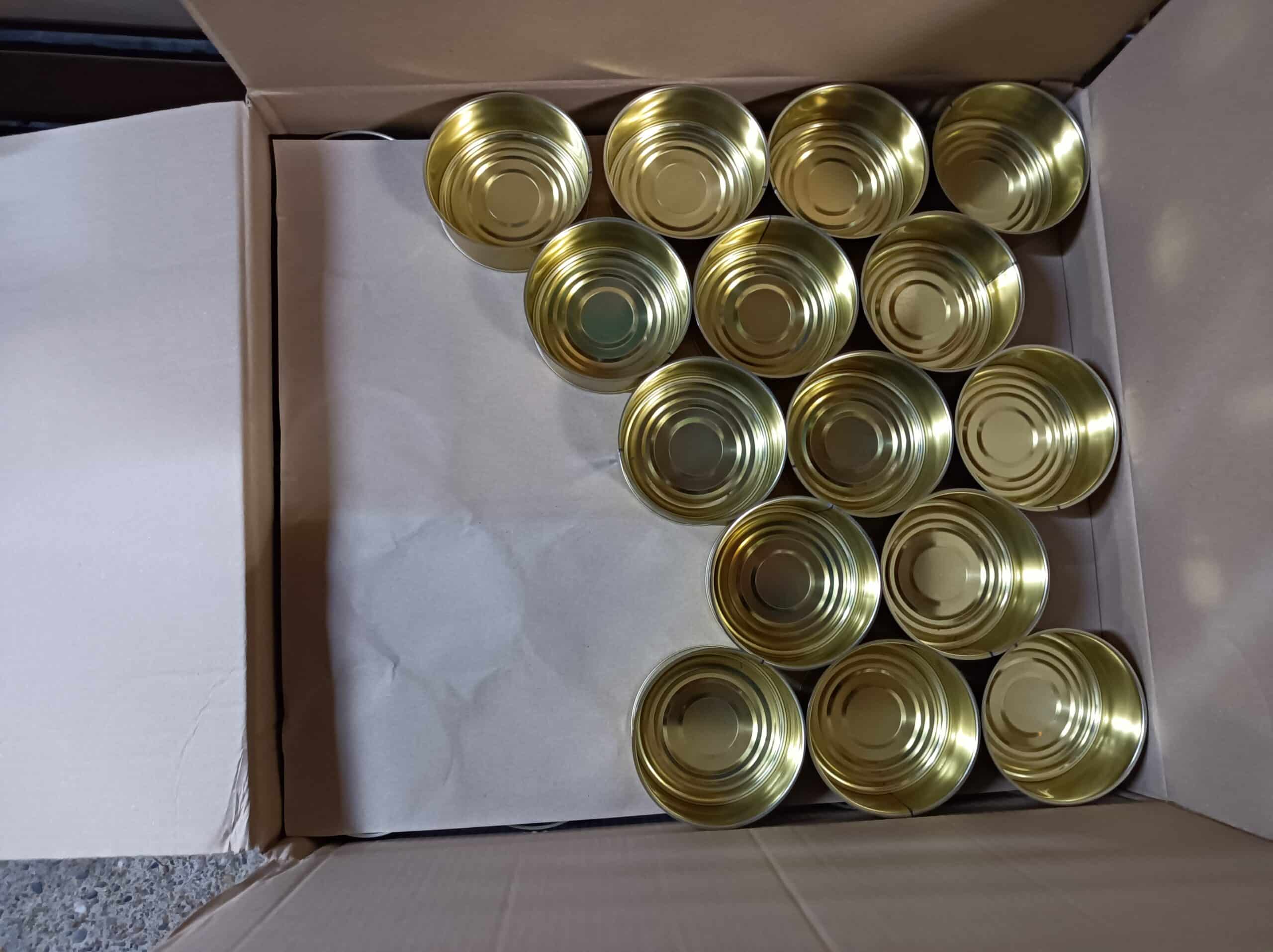 Nombreuses boîtes de conserve vides dorées, neuves et brillantes, disposées dans une boîte en carton ouverte. Les fonds sont striés.