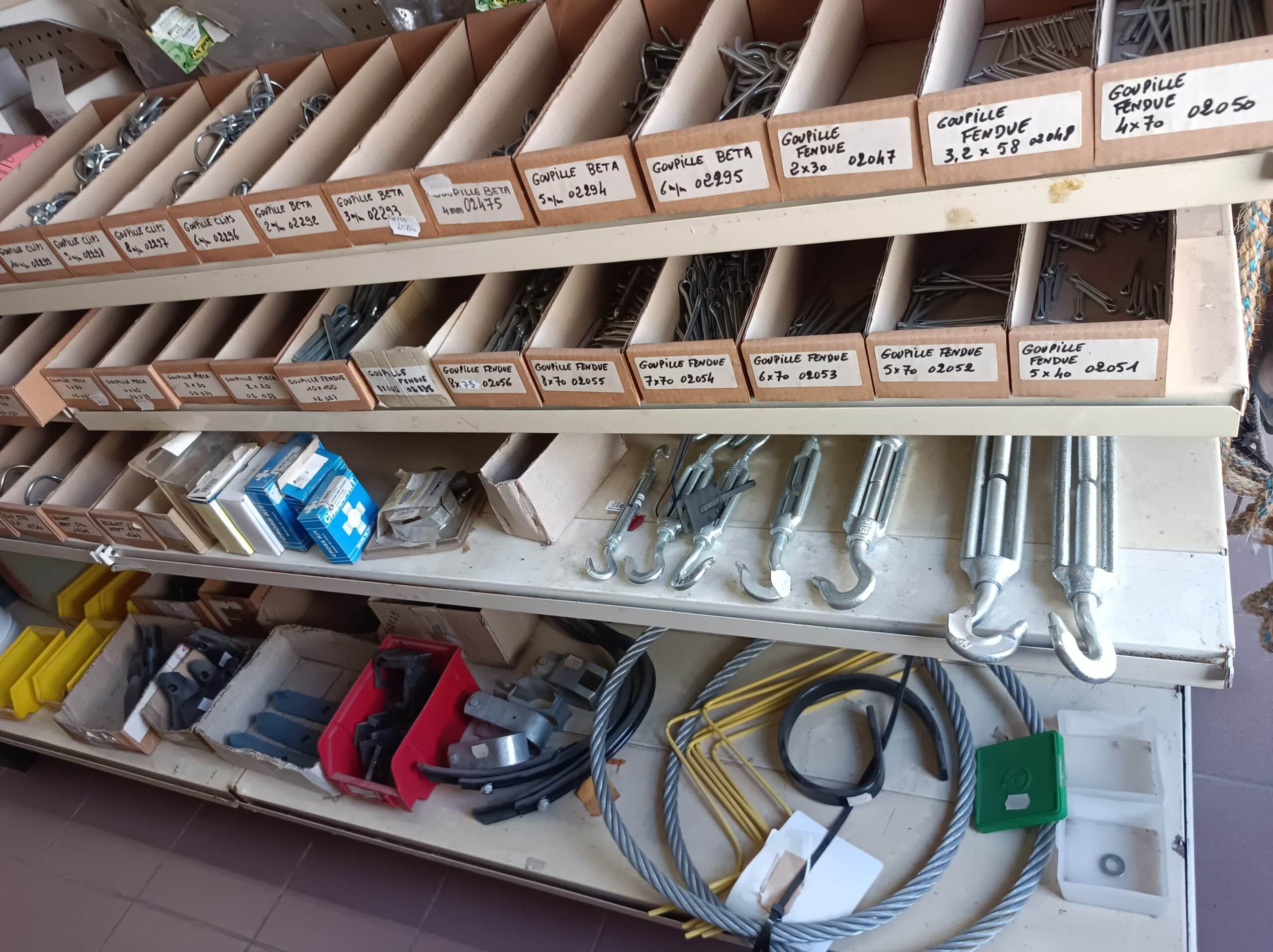 Étagères de quincaillerie avec des boîtes étiquetées "goupille clips", "fendue", "bêta" de diverses tailles, tendeurs et câbles. Stock organisé.