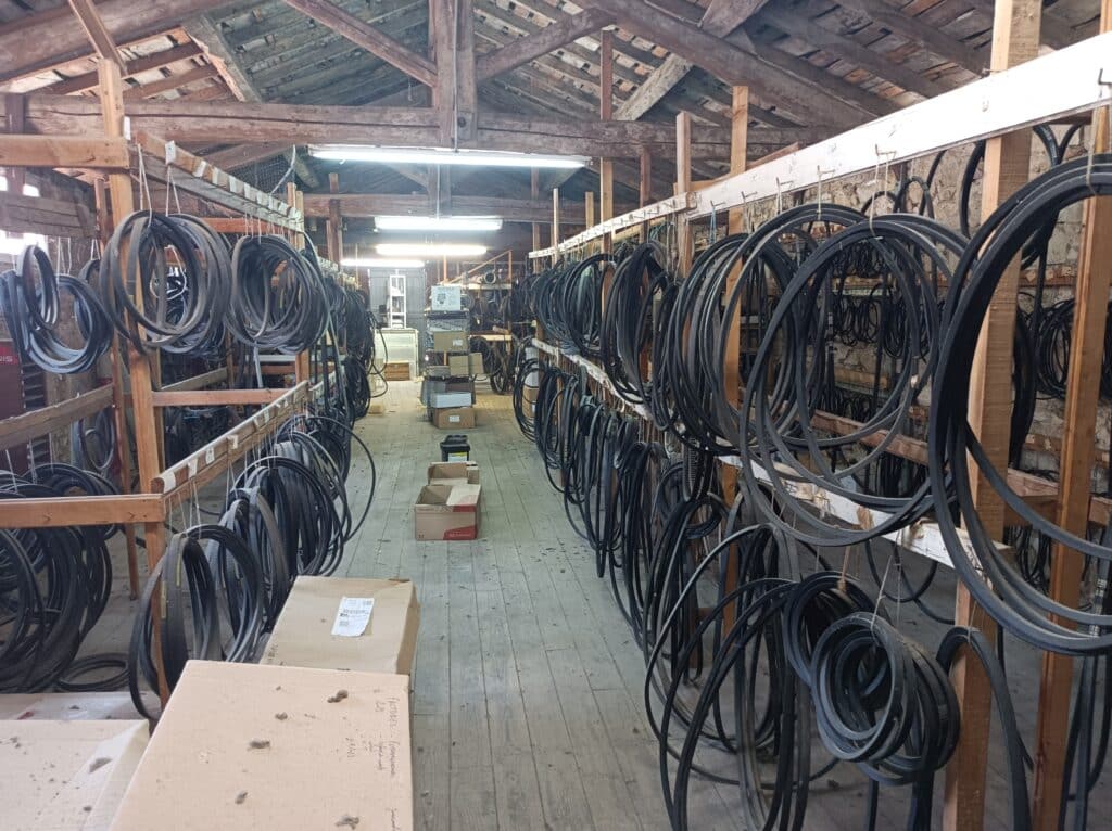 Allée de stockage avec des centaines de courroies noires sur racks en bois sous poutres apparentes. Boîtes Kramp au sol. Texte : FACTURES.